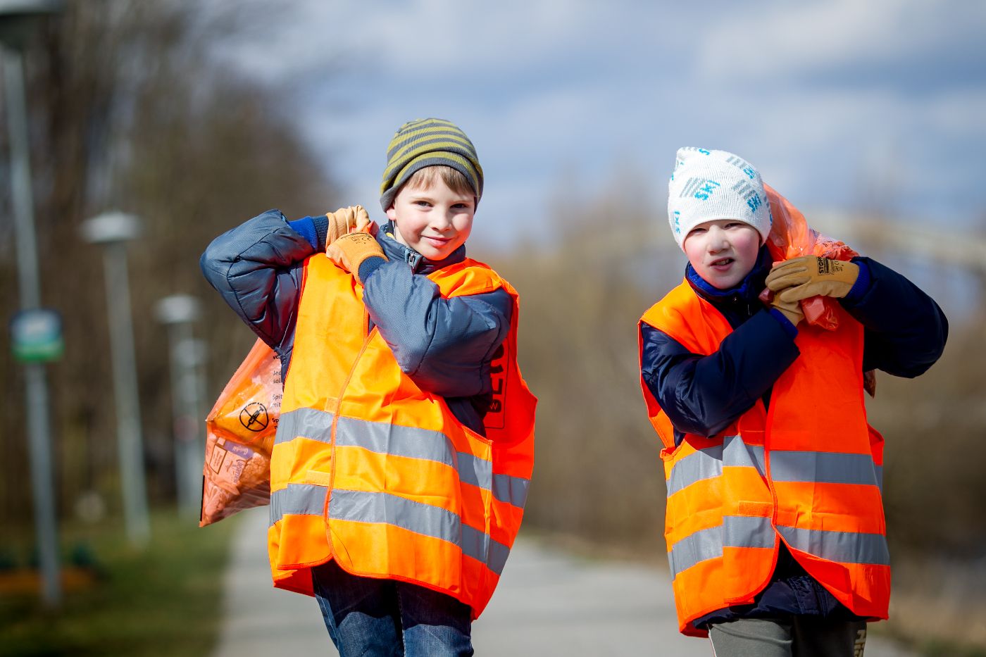 Auf dem Bild sind zwei junge Buben in Warnweste mit blauen Jacken gekleidet abgebildet. Beide haben einen orangen Sack mit Abfllen ber die Schulter geworfen und blicken in die Kamera. 