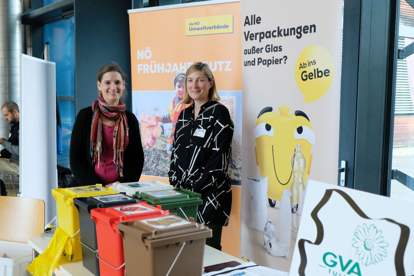 Auf dem Bild sind zwei Frauen zu sehen, die in die Kamera blicken. Im Vordergrund sind kleine Kbel in unterschiedlichen Farben fr die jeweilige Zuordnung der Abfalltrennung zu erkennen. 